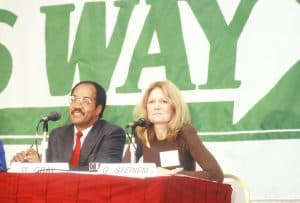 Gloria Steinem sits at a table with a microphone in front of her. The words 'WAY' from the WOMEN'S WAY logo are visible beside her. She is sitting next to W. Gray.