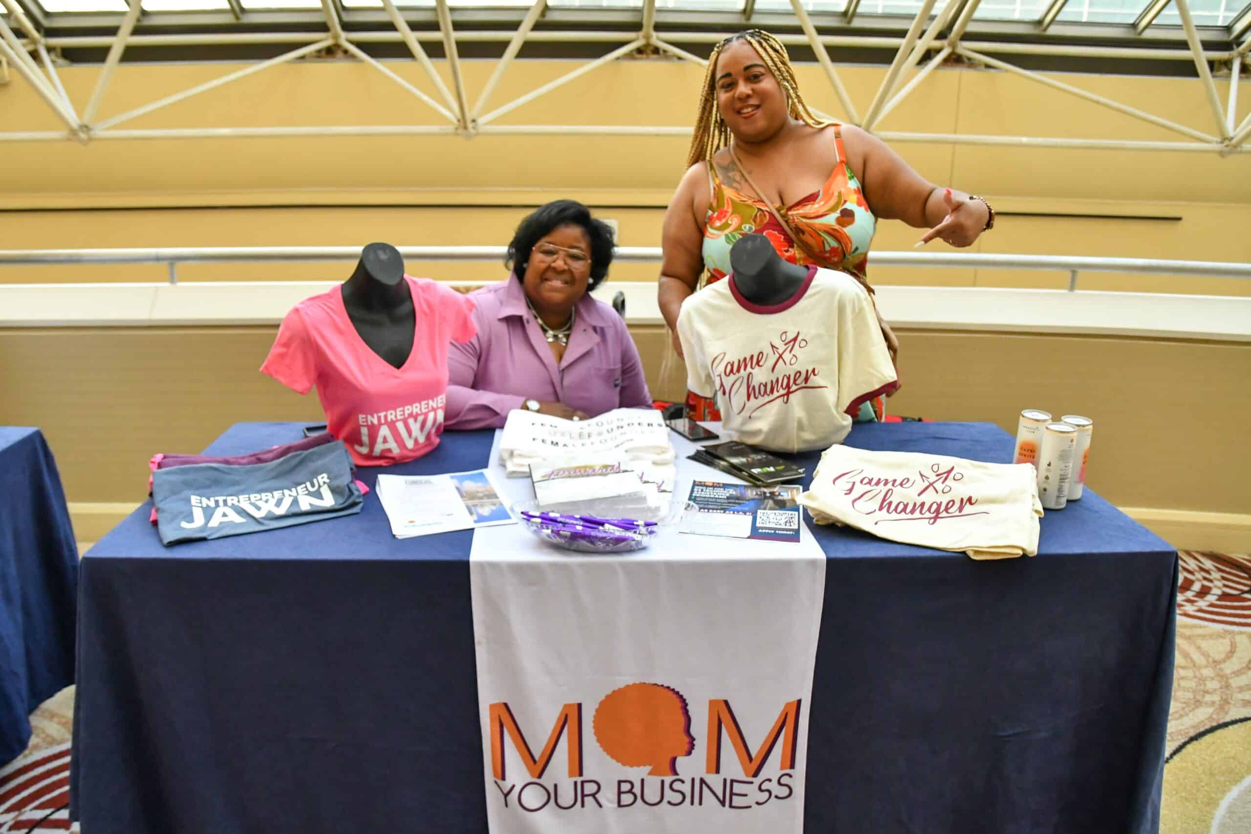 Two Black women business owners sit at a table with a Mom Your Business Banner. There is other branded merchandise on the table to promote their business.
